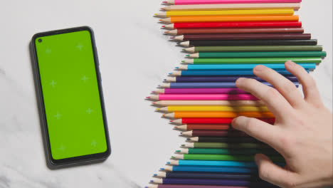 Coloured-Pencils-In-A-Line-On-Marble-Background-With-Person-Choosing-Red-One-And-Green-Screen-Mobile-Phone-1
