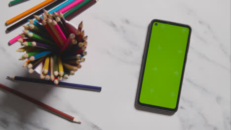 Overhead-Shot-Of-Multi-Coloured-Pencils-In-Pot-And-Green-Screen-Phone-On-Marble-Background