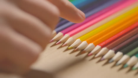Studio-Shot-Of-Multi-Coloured-Pencils-In-A-Line-On-Wooden-Background-With-Hand-Choosing