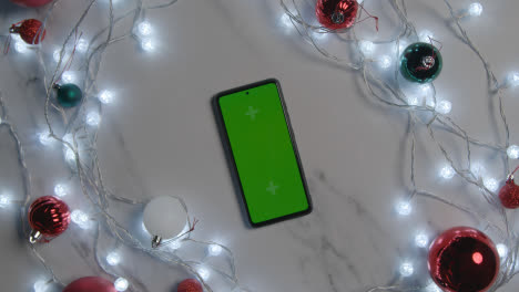 Overhead-Shot-Of-Revolving-Green-Screen-Mobile-Phone-With-Christmas-Decorations-And-Lights