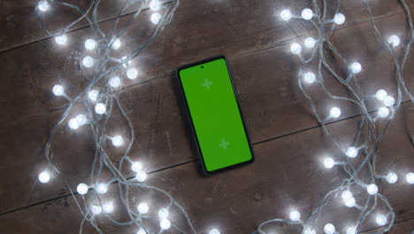 Overhead-Aufnahme-Eines-Sich-Drehenden-Greenscreen-Mobiltelefons-Mit-Weihnachtsdekorationen-Und-Lichtern-2