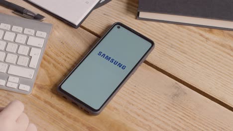 Mobile-Phone-On-Desk-With-Screen-Showing-Brand-Logos-For-Apple-And-Samsung