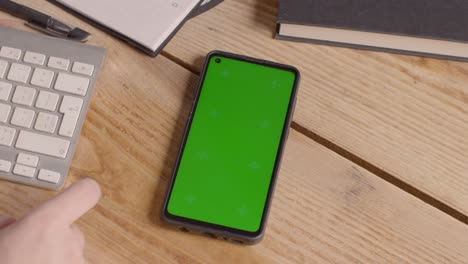 Nahaufnahme-Eines-Mobiltelefons-Mit-Grünem-Bildschirm-Auf-Dem-Schreibtisch,-Wobei-Die-Hand-über-Den-Bildschirm-Scrollt
