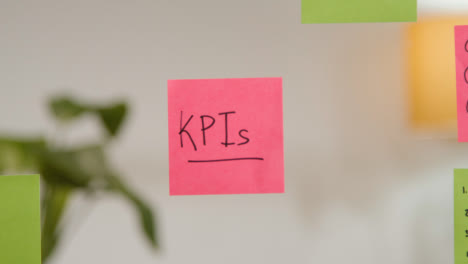 Close-Up-Of-Woman-Writing-KPIs-Onto-Sticky-Note-Stuck-To-Transparent-Screen-In-Office