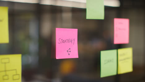 Close-Up-Of-Woman-Putting-Sticky-Note-With-Strategy-Written-On-It-Onto-Transparent-Screen-In-Office-1