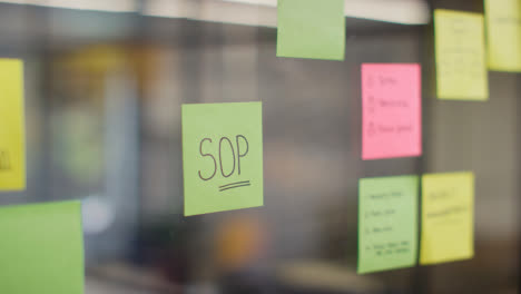 Close-Up-Of-Woman-Putting-Sticky-Note-With-SOP-Written-On-It-Onto-Transparent-Screen-In-Office-1