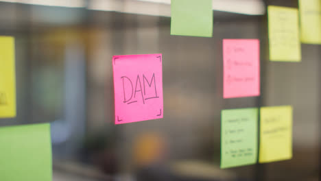Close-Up-Of-Woman-Putting-Sticky-Note-With-DAM-Written-On-It-Onto-Transparent-Screen-In-Office