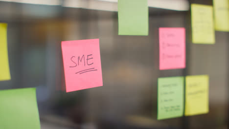 Close-Up-Of-Woman-Putting-Sticky-Note-With-SME-Written-On-It-Onto-Transparent-Screen-In-Office-1