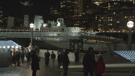 Busy-Christmas-Market-Stalls-Looking-Towards-HMS-Belfast-On-London-South-Bank-At-Night