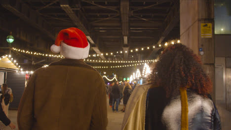 Couple-Celebrating-Christmas-Walking-Through-Market-Stalls-On-London-South-Bank-At-Night