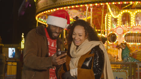 Pareja-Celebrando-La-Navidad-De-Pie-Junto-A-La-Rotonda-Del-Recinto-Ferial-En-El-Banco-Sur-De-Londres-Por-La-Noche-Con-Teléfono-Móvil