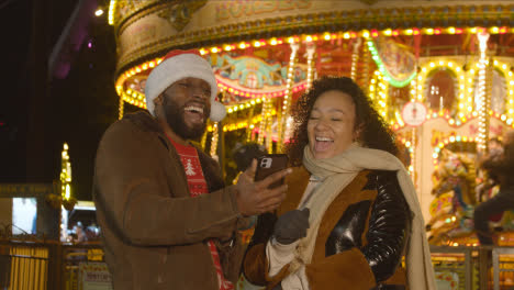 Couple-Celebrating-Christmas-Standing-By-Fairground-Roundabout-On-London-South-Bank-At-Night-With-Mobile-Phone-2
