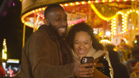 Couple-Standing-By-Fairground-Roundabout-On-London-South-Bank-Making-Video-Call-On-Mobile-Phone-