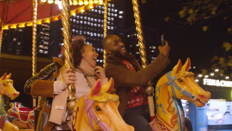 Couple-Having-Fun-Riding-On-Fairground-Roundabout-On-London-South-Bank-At-Night-With-Mobile-Phone