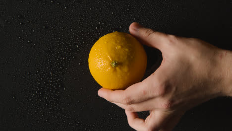 Overhead-Studio-Shot-Of-Hand-Picking-Up-Orange-With-Water-Droplets-Revolving-Against-Black-Background