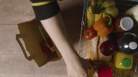 Overhead-Shot-Of-Person-Packing-Basic-Fresh-Food-Items-From-Supermarket-Wire-Shopping-Basket-Into-Paper-Bag