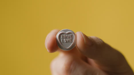 Close-Up-Of-Hand-Holding-Heart-Candy-With-True-Love-Message-On-Yellow-Background