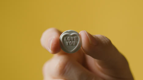 Close-Up-Of-Hand-Holding-Heart-Candy-With-Love-You-Message-On-Yellow-Background
