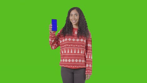 Studio-Portrait-Of-Woman-Wearing-Christmas-Jumper-Holding-Up-Mobile-Phone-Against-Green-Screen-1