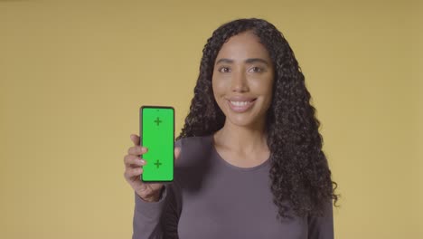 Studio-Portrait-Of-Smiling-Woman-Holding-Up-Mobile-Phone-With-Green-Screen