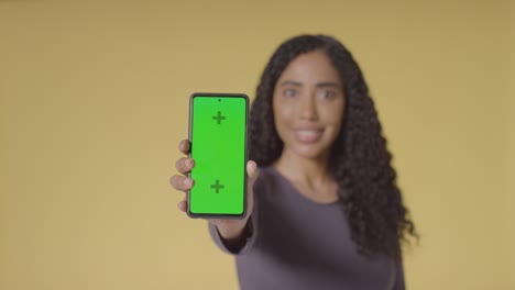 Studio-Portrait-Of-Smiling-Woman-Holding-Up-Mobile-Phone-With-Green-Screen