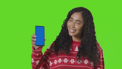 Studio-Portrait-Of-Woman-Wearing-Christmas-Jumper-Holding-Up-Mobile-Phone-Against-Green-Screen