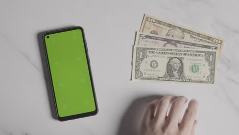 Overhead-Currency-Shot-Of-Person-Tapping-Fingers-Next-To-Dollar-Bills-And-Green-Screen-Mobile-Phone