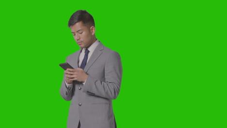 Studio-Shot-Of-Businessman-In-Suit-Looking-At-Mobile-Phone-Against-Green-Screen-