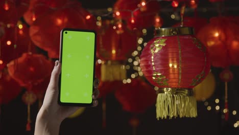 Mano-Sosteniendo-Un-Teléfono-Móvil-De-Pantalla-Verde-Con-Linternas-Chinas-Colgadas-En-El-Fondo-2