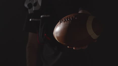 Close-Up-Studio-Shot-Of-American-Football-Player-Holding-Out-Ball-With-Low-Key-Lighting-