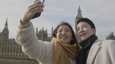 Joven-Pareja-Asiática-De-Vacaciones-Posando-Para-Selfie-Frente-A-Las-Casas-Del-Parlamento-En-Londres,-Reino-Unido