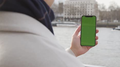Primer-Plano-De-Un-Hombre-Que-Sostiene-Un-Teléfono-Móvil-Con-Pantalla-Verde-Mirando-Hacia-El-Río-Támesis-Y-El-Terraplén-En-Londres,-Reino-Unido-1