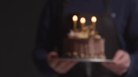Studio-Shot-Of-Person-Carrying-Decorated-Chocolate-Birthday-Celebration-Cake-With-Lit-Candles-Against-Black-Background