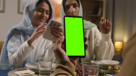 Mann-Blickt-Auf-Ein-Mobiltelefon-Mit-Grünem-Bildschirm,-Sitzt-Zu-Hause-Am-Muslimischen-Familientisch-Und-Isst-Iftar-Mahlzeit-Und-Bricht-Während-Des-Ramadan-Täglich-Das-Fasten