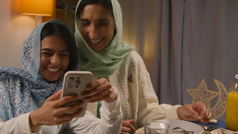 Mutter-Und-Tochter-Lachen-über-Ihr-Mobiltelefon,-Sitzen-Zu-Hause-Am-Muslimischen-Familientisch-Und-Essen-Iftar-Mahlzeit-Und-Brechen-Während-Ramadan-1-Täglich-Das-Fasten