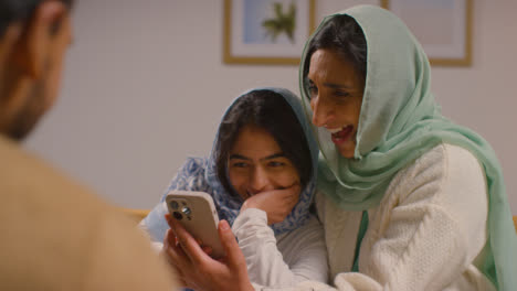 Muslimische-Mutter-Und-Tochter-Lachen-über-Das-Mobiltelefon-Und-Sitzen-Zu-Hause-Am-Muslimischen-Familientisch