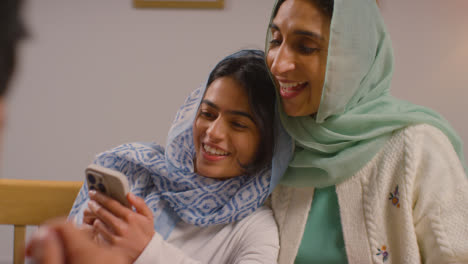 Muslimische-Mutter-Und-Tochter-Lachen-über-Ihr-Mobiltelefon-Und-Sitzen-Zu-Hause-Am-Muslimischen-Familientisch-1
