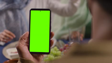Mann-Blickt-Auf-Ein-Mobiltelefon-Mit-Grünem-Bildschirm,-Sitzt-Zu-Hause-Am-Tisch-Einer-Muslimischen-Familie-Und-Isst-Eine-Mahlzeit