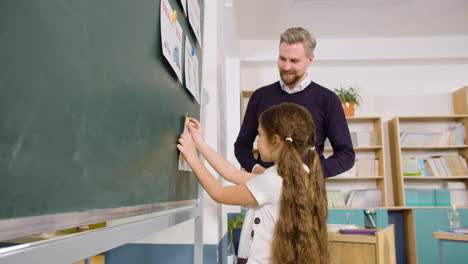 Side-View-Of-A-Teacher-In-Front-Of-The-Blackboard-In-English-Classroom,-Then-A-Female-Student-Stands-Up-And-Sticks-A-Paper-With-The-Words-'Happy'-And-'sad'-On-Blackboard