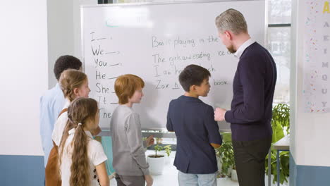 Multiethnic-Students-Group-And-Teacher-Standing-In-Front-Of-The-Blackboard-While-A-Ginger-Student-Helps-An-Classmate-To-Point-Out-The-Verb-To-Be
