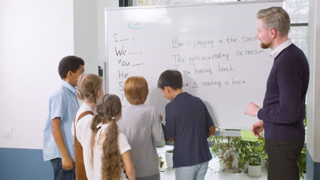 Multiethnic-Students-Group-And-Teacher-Standing-In-Front-Of-The-Blackboard-While-A-Student-Point-Out-The-Verb-To-Be