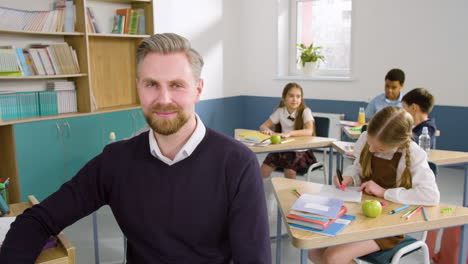 Lehrerin-Schaut-Im-Englischen-Klassenzimmer-In-Die-Kamera,-Im-Hintergrund-Sitzen-Ihre-Schüler-An-Schreibtischen