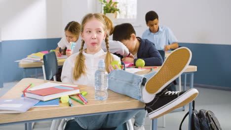 Female-Student-Sitting-At-The-Desk-With-Her-Legs-On-The-Table-In-English-Classroom-While-Eating-An-Apple