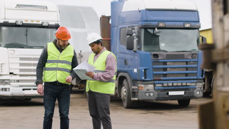 Distant-View-Of-A-Boss-And-Worker-Wearing-Vests-And-Safety-Helmets-Organizing-A-Truck-Fleet-In-A-Logistics-Park-While-They-Consulting-A-Document