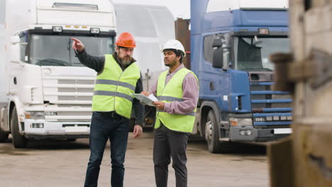 Distant-View-Of-A-Boss-And-Worker-Wearing-Vests-And-Safety-Helmets-Organizing-A-Truck-Fleet-In-A-Logistics-Park-While-They-Consulting-A-Document-1