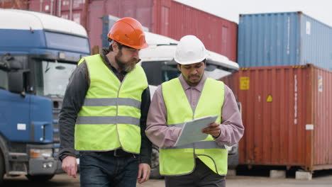 Boss-And-Worker-Wearing-Vests-And-Safety-Helmets-Organizing-A-Truck-Fleet-In-A-Logistics-Park-While-They-Consulting-A-Document-And-Walking