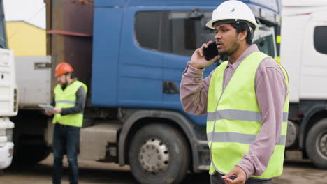 Worker-Wearing-Vest-And-Safety-Helmet-Organizing-A-Truck-Fleet-In-A-Logistics-Park-While-Talking-On-The-Phone