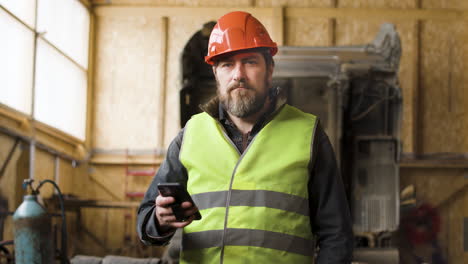 Worker-Wearing-Vest-And-Safety-Helmet-Holding-A-Smartphone-And-Looking-At-Camera-In-A-Logistics-Park