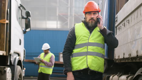 Worker-Wearing-Vest-And-Safety-Helmet-Organizing-A-Truck-Fleet-In-A-Logistics-Park-While-Talking-On-The-Phone-1