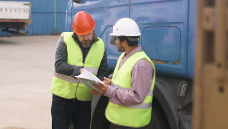Boss-And-Worker-Wearing-Vests-And-Safety-Helmets-Organizing-A-Truck-Fleet-In-A-Logistics-Park-While-They-Consulting-A-Document-9
